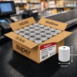 Open box of silver‑wrapped Super thermal paper rolls on a POS counter beside an 80 mm roll